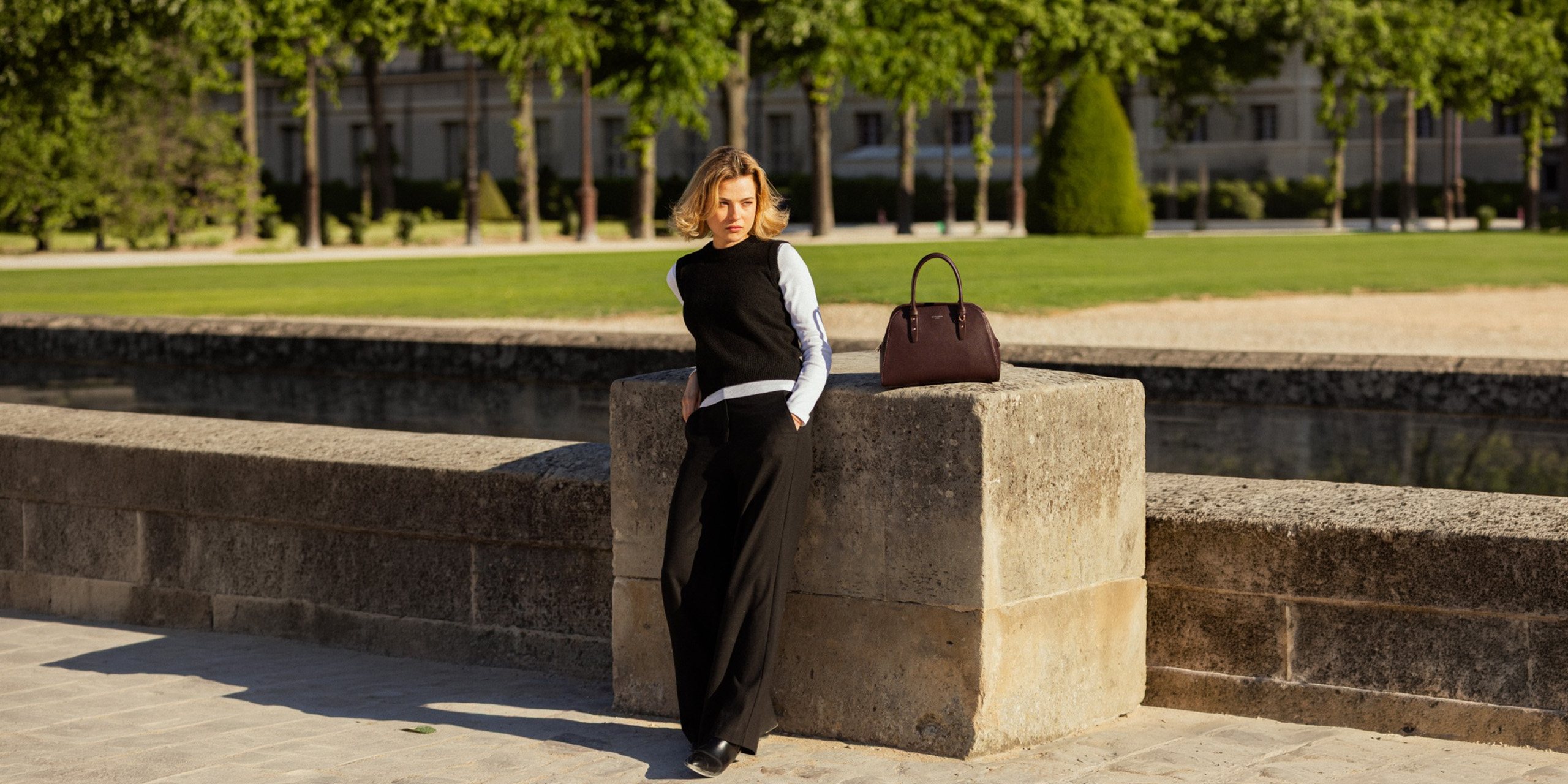 Mujer recargada en un muro de piedra en París junto a un bolso de mano David Jones color vino